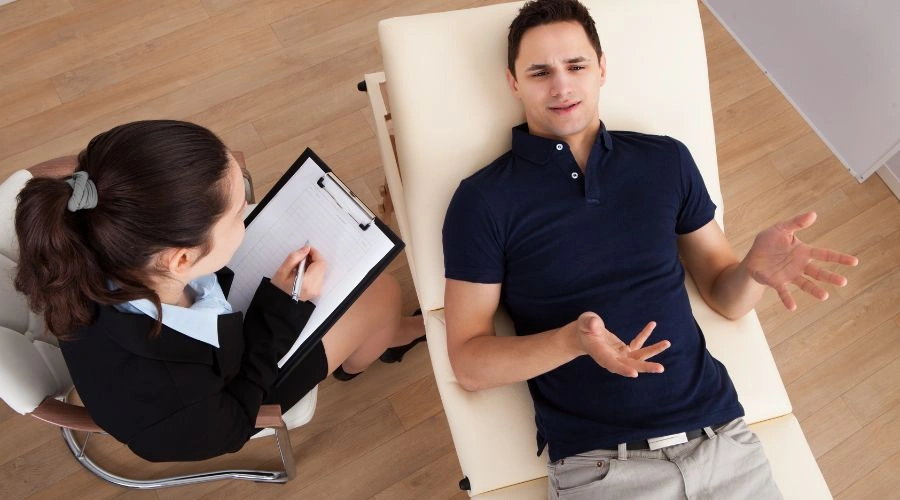 Patient laying on a table talking to counselor interviewing him who is working to Create the report that summarizes Patient findings