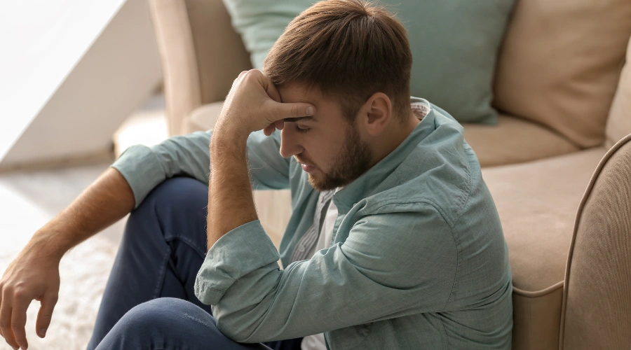 Image of a man sitting on the floor in front of a sofa in a pensive manner, holding his hand on his forehead